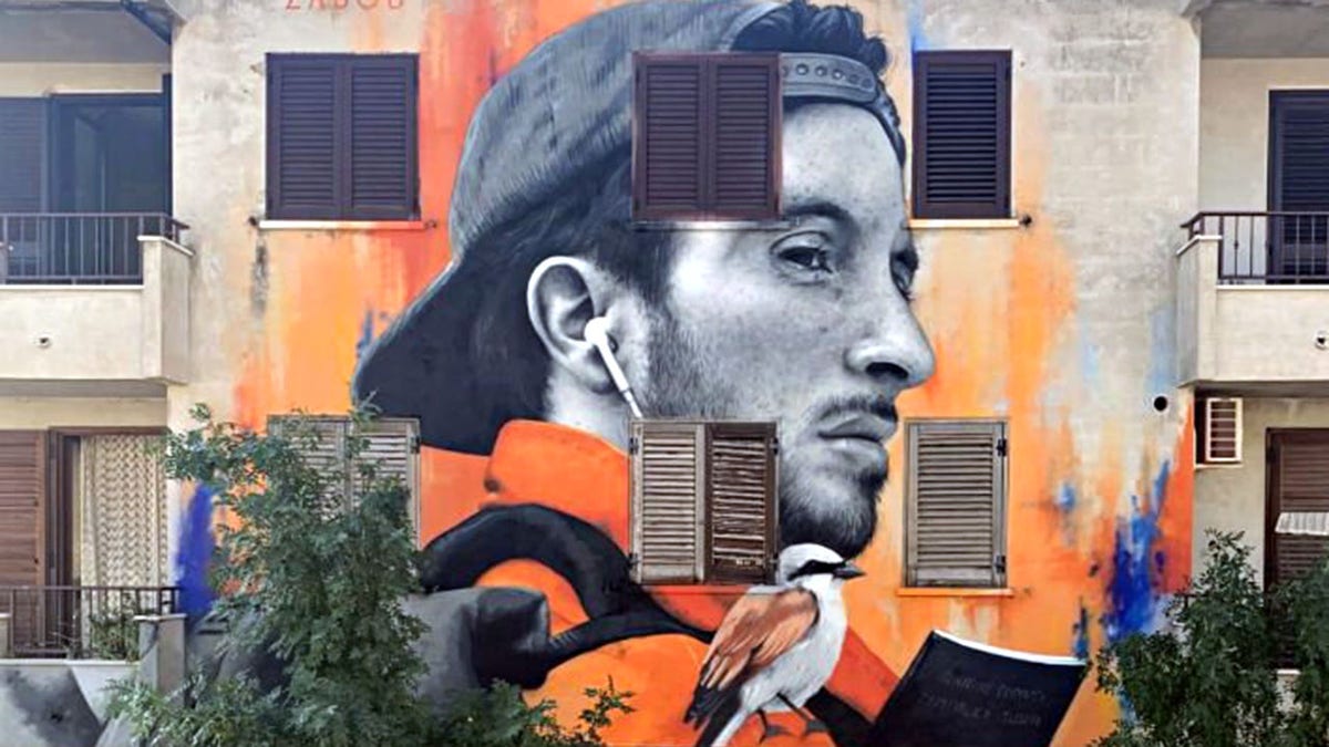 mural of a man with airpods in and an orange sweater with a bird on his shoulder as he turns his head to the right, painted on the side of a building in Stornara, Italy