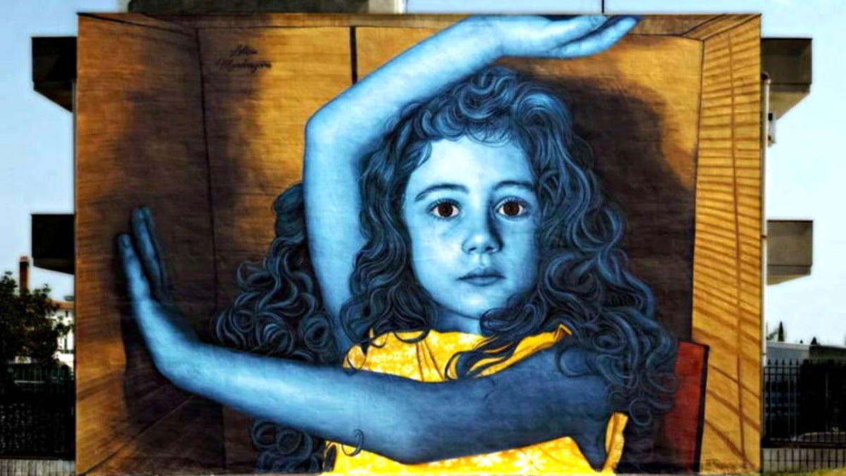 Mural of a little girl on the side of a building wall in Stornara, Italy. In the mural, her skin is tinted blue, she has wavy hair and she is wearing a yellow dress, with one hand over her head and one across her body, seemingly within a box