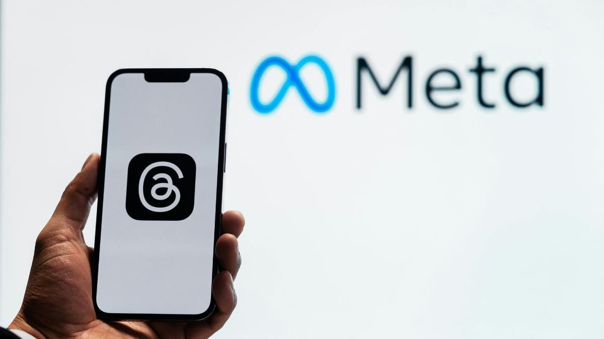 A smartphone displaying the logo of the Threads app is held in front of a screen displaying the Meta logo.