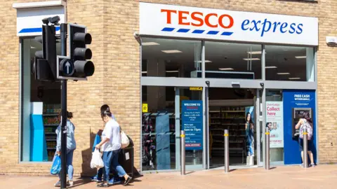 Getty Images Three people walk past a Tesco Express carrying plastic shopping bags, while someone else uses a Tesco ATM at the front of the store.