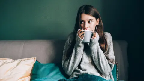 Getty Images Woman in a jumper under a blanket with a mug of tea. She is hunching her shoulders as if feeling cold
