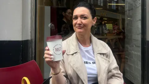 Mimoza Emsa with dark brown hair in a ponytail wearing a beige jacket over  a white t shirt sitting outside Pret holding a takeaway coffee cup