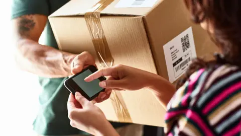 Getty Images A delivery man in a green t-shirt hands a brown cardboard box to a woman with brown hair, wearing a stripy top. The delivery man is holding a phone up for the woman to sign for the package.