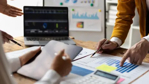 getty Work colleagues reviewing charts and graphs on a desk in an office. One is standing up pointing at a chart, while another person is sitting down reading a tablet.