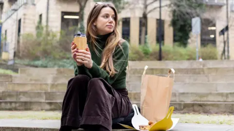 Getty Images A young woman holding a reusable coffee cup sits on an outdoor concrete step with a brown paper bag and an open box containing food.