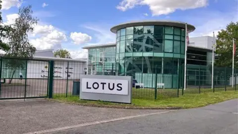 Martin Giles/BBC The outside of the Lotus building in Hethel, near Wymondham. The building is a curved glass building with a sign outside the front which says LOTUS in black letters.