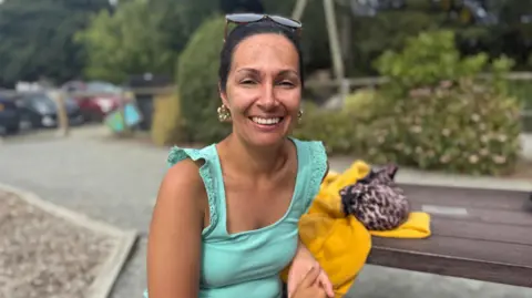 Women in her 30s in a blue top leant on a yellow jumper. She has brunette hair up in a claw clip and big gold earrings