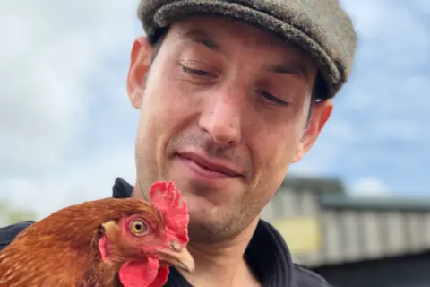 Lewis Clare Lewis is wearing a dark sweater with a tweed flat cap on his head. He holds a gingery-brown chicken close to his chest while standing outside a barn, with a partly cloudy sky and part of a building visible in the background.