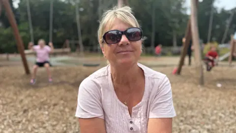 a woman in her late 50's wearing a light pink cotton top. She wears round sunglasses and has blonde hair up in a ponytail