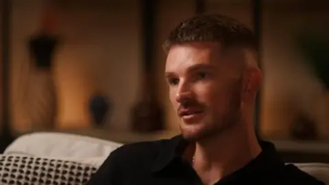 We Need To Talk Adam Frisby, a white man with short, fair hair and a short beard. He is sitting on a sofa being interviewed for a podcast