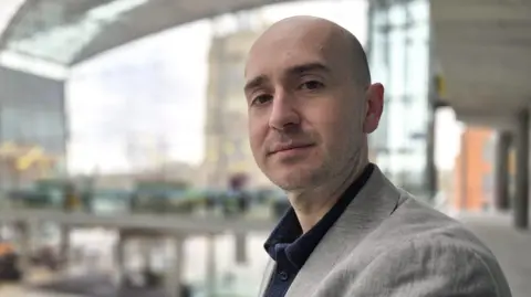 Paul Moseley/BBC Ben Goldsborough who is standing inside The Forum in Norwich. He is looking directly at the camera and is wearing a grey suit jacket and dark shirt. 