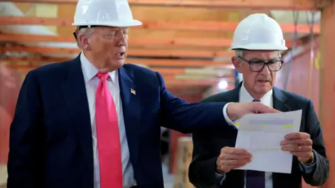 Chip Somodevilla/ Getty Images US President Donald Trump and Federal Reserve Chair Jerome Powell tour the Federal Reserve’s headquarters renovation project on July 24, 2025. Both men are wearing white hard hats, and suits, as Trump points at a piece of paper Powell is holding, which both men are looking at.