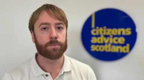 BBC David Hilferty, director of impact at Citizens Advice Scotland, sits in front of a sign displaying the charity's logo. He has brown hair and a brown beard and wears a white shirt.