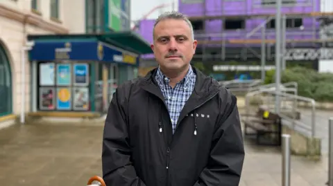 BBC A man, looking directly at the camera wearing a blue and white checkered top and black coat. Behind him is a building with scaffolding. The ground is pavement.