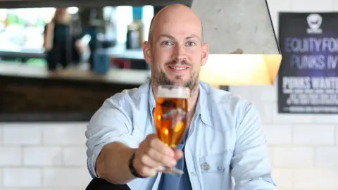 Getty Images James Watt holding a glass of beer to the camera. He is sitting in a bar wearing a light blue shirt and a blue t-shirt.