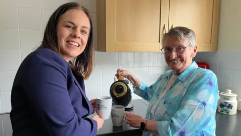 BBC Kate Forbes, a dark-haired woman in a blue suit, holds a mug while Muriel Allison, who has grey hair and is wearing a light blue blouse, pours water from a kettle.