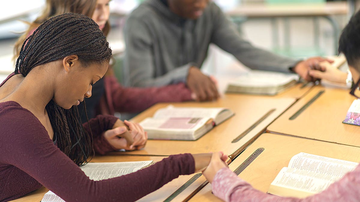 College students seated at desks holding hands in prayer.