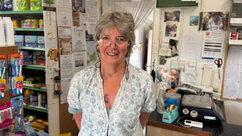 BBC A lady with grey short hair standing in a shop. there are toothbrushes, petfood and a till in the background.