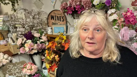 John Devine/BBC Andrea Moat, 60, has shoulder-length blonde hair, blue eyes and has a pair of glasses resting on her head. She has a thick black woollen jumper on and behind her are cut flowers in vases and various ornamental signs.
