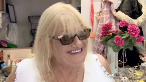 Lynne Russell has medium-length blonde hair with a fringe. She's wearing tortoiseshell sunglasses, a white t-shirt and is smiling inside the Helping Hands shop standing next to some pink flowers.. In the background, various items of clothing and ornaments can be seen. 