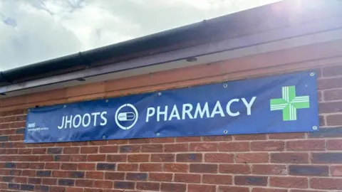BBC A Blue NHS branded banner which reads "Jhoots Pharmacy" is shown stuck to a brick wall. The NHS symbol can be seen in the far left, with the pharmacy symbol in the far right.