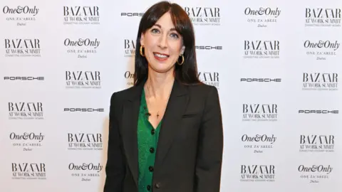 Dave Bennett/Getty Images Samanthan Cameron wearing a green cardigan under a black blazer, at a magazine event with the promotional board behind her. She is smiling and wearing gold hoop earrings.