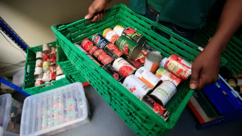 PA Media Someone carries a green crate filled with cans of tomatoes.  Another crate and a  clear plastic container has other items in it.