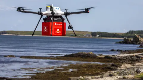 Skyports/Royal Mail A white Skyports drones carries a red box with the Royal Mail logo, over a beach with the sea in the background.