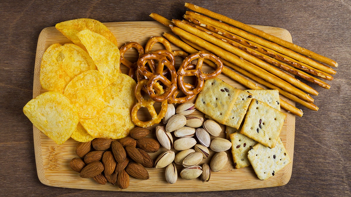 Assortment snacks including almonds, pistachios, crackers, potato chips and mini pretzels seen on a wooding cutting board.