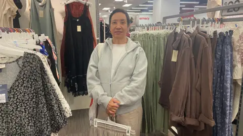 Danielle Kaye/BBC Nancy Garcia, who works in the publishing and gifts industry, shops for clothing at a Target location in Manhattan on 10 Sept 2025. She is surrounded by dresses and jackets on hangers and is holding some clothes.