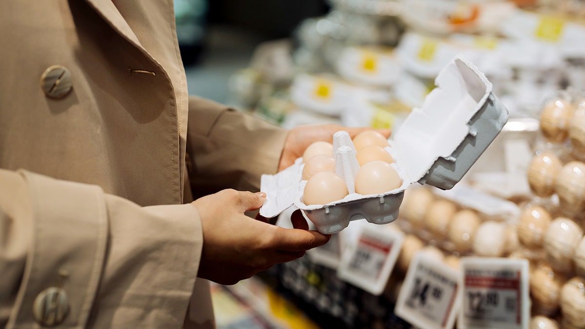 A woman wearing a brown jacket holds a carton of organic eggs at a grocery store.