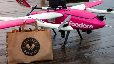 Aviant The foodora drone is pink and white with rotors on mounts attached to the wings - a third propeller is on the back. In the foreground is a brown paper bag from Bastard Burgers.
