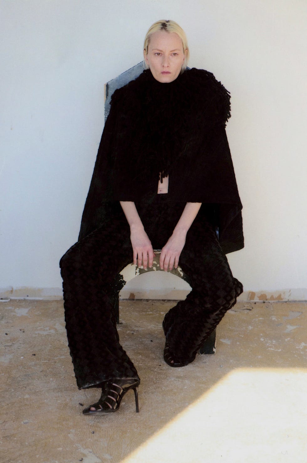 fashion model wearing dark textured outfit sitting on a chair against a blank wall