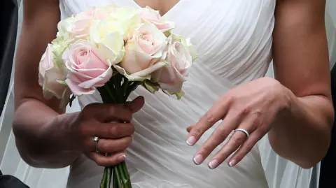 BBC A bride pictured from the neck down. She is wearing a white wedding dress and holding a bouquet of white and pink roses. She has a French manicure and is wearing an engagement ring on her left hand and a wedding band on her right hand