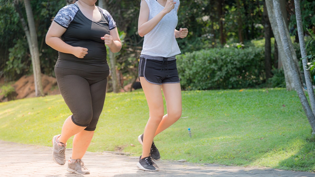 One heavy and one thin woman, seen only from necks down, jogging on outdoor path together.